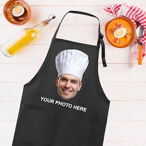 May include: A black apron with a large pocket on the front. The apron has a white chef's hat with a face printed on it. The text "YOUR PHOTO HERE" is printed below the face.