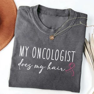 May include: Gray t-shirt with the text "My Oncologist does my hair" in white lettering and a pink ribbon graphic.
