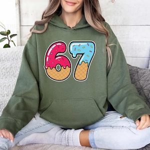 May include: An olive green hooded sweatshirt featuring a colorful graphic of the number "67" designed to look like donuts. The "6" is pink with white frosting and the "7" is blue with sprinkles. The numbers have a waffle cone design.