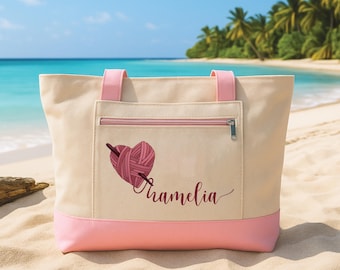 May include: A beige tote bag with pink accents, featuring a heart-shaped yarn design and the name "Tramelia" in cursive. The bag has a zippered pocket and pink handles, set against a beach backdrop.