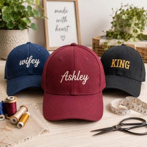 May include: Three baseball caps in navy blue, maroon, and black. The navy cap features the white embroidered text 'wifey', the maroon cap displays the white embroidered name 'Ashley', and the black cap has the yellow embroidered text 'KING'.