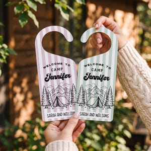 May include: Two door hangers with vertical stripes in light purple and light green. Each features a black line drawing of a tent, trees, and mountains. Text reads: Welcome to Camp Jennifer, Sarah and William. The hangers have a curved hook top.