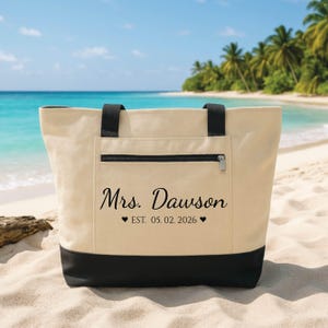 May include: A beige canvas tote bag with black accents, a zippered pocket, and black handles. The bag features the text "Mrs. Dawson" with the date "05.02.2026" printed below. The bag is on a sandy beach with a turquoise ocean and palm trees in the background.