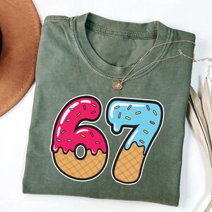 May include: An olive green t-shirt with a large graphic of the number 67, designed to look like a donut and ice cream cone. The number 6 is pink with sprinkles, and the 7 is blue with sprinkles. A gold necklace is draped across the shirt.