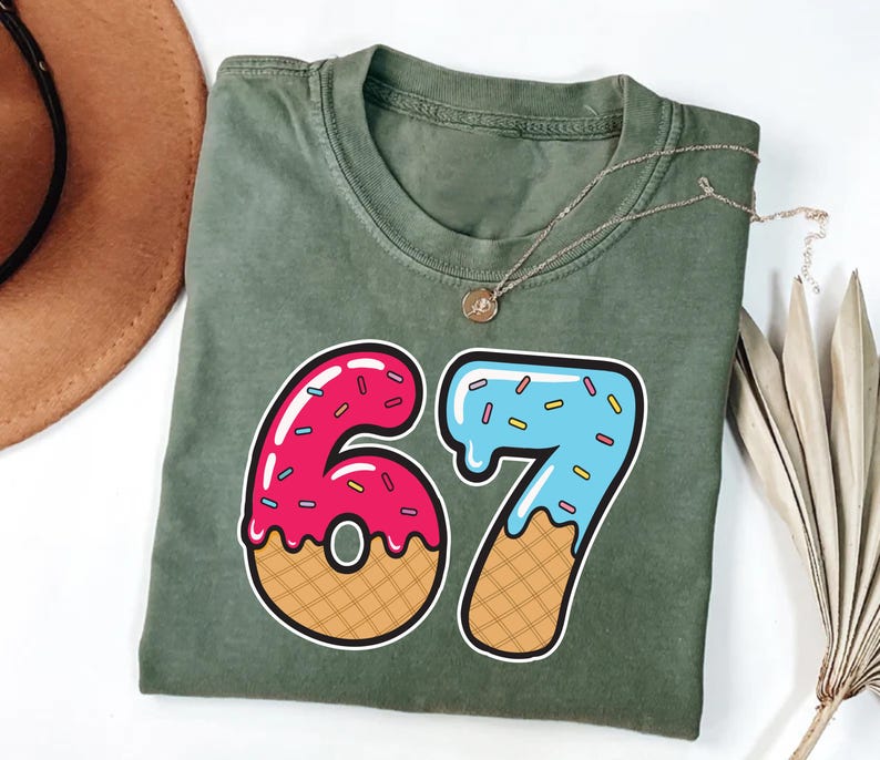 May include: An olive green t-shirt with a large graphic of the number 67, designed to look like a donut and ice cream cone. The number 6 is pink with sprinkles, and the 7 is blue with sprinkles. A gold necklace is draped across the shirt.