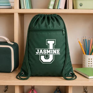 May include: A dark green drawstring backpack featuring a white graphic of a large letter J with the name JASMINE printed horizontally across the center. The bag includes a horizontal zippered pocket on the front and thick green cord straps.