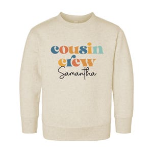 May include: A beige sweatshirt with the text "cousin crew Samantha" in colorful retro-style lettering.