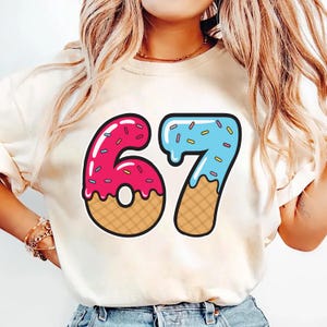 May include: Cream-colored t-shirt featuring the number 67 in a playful, dessert-themed design. The number 6 is pink with sprinkles, and the number 7 is blue with sprinkles. Both numbers have a waffle cone base.