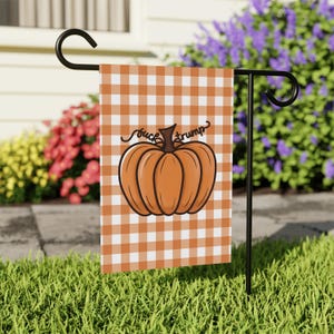 May include: A garden flag with a pumpkin design and the text "fuck trump" on a white and orange checkered background. The flag is displayed on a black metal stand in a grassy yard.