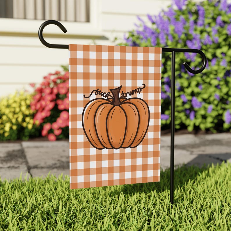May include: A garden flag with a pumpkin design and the text "fuck trump" on a white and orange checkered background. The flag is displayed on a black metal stand in a grassy yard.