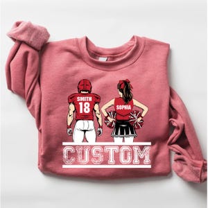 May include: A dusty rose sweatshirt featuring a graphic of a football player and a cheerleader. The football player's jersey reads "SMITH 18", and the cheerleader's top says "SOPHIA". The word "CUSTOM" is printed in a white, distressed font.