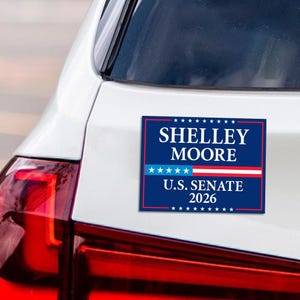 May include: A blue and white car sticker with the text "SHELLEY MOORE U.S. SENATE 2026". The sticker is attached to the back of a white car, with the red taillight visible on the left.