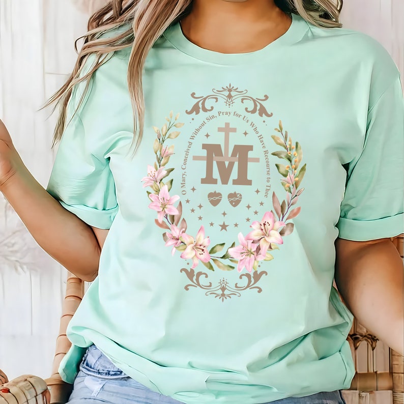 Op de afbeelding: Een lichtblauw t-shirt met een bloemenkransontwerp met een bruine afbeelding van de Maagd Maria. De tekst luidt "O Maria, ontvangen zonder zonde, bid voor ons die onze toevlucht tot u nemen".
