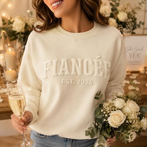 May include: A cream-colored crewneck sweatshirt featuring the word FIANCÉE in raised, tonal lettering across the chest, with the text EST. 2026 printed directly below in a matching font. The garment has long sleeves and a ribbed neckline.