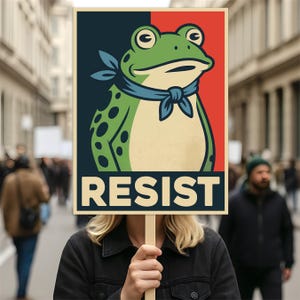 May include: A poster with a frog illustration wearing a blue scarf, set against a red and navy background. The word "RESIST" is printed in bold, cream-colored letters. The poster is held up in a crowd.