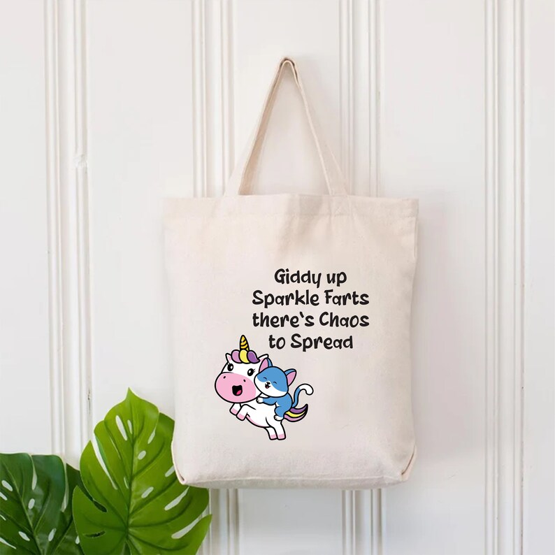 May include: A white canvas tote bag with a cartoon unicorn and cat. The text on the bag reads "Giddy up Sparkle Farts there's Chaos to Spread".
