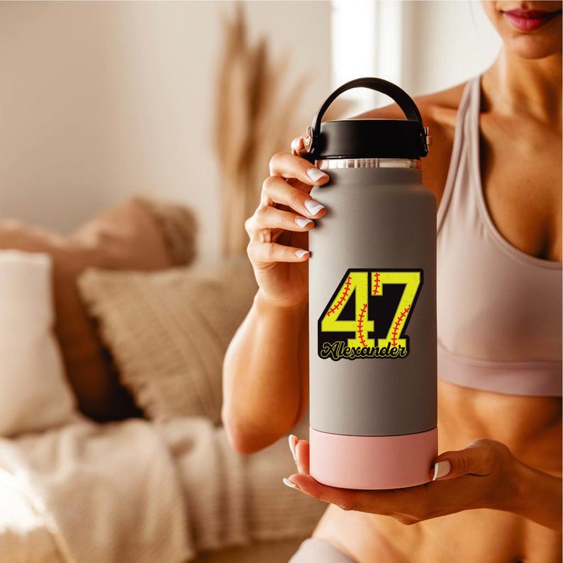 May include: A gray water bottle with a black handle and a pink base. The bottle features a yellow and black graphic with the number 47 and the name "Alexander" in a baseball-themed design. The bottle is held by a person.