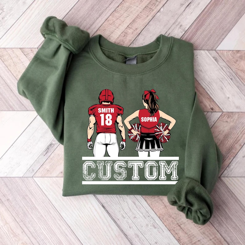 May include: Green sweatshirt with a graphic of a football player and cheerleader. The football player wears a red helmet and jersey with the name "Smith" and the number "18." The cheerleader wears a red top and black skirt with the name "Sophia." The word "CUSTOM" is below.