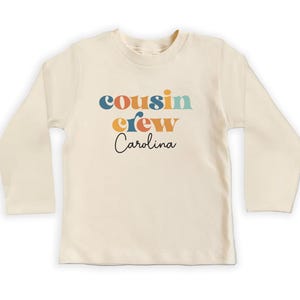 May include: A long-sleeved, cream-colored t-shirt with the text "cousin crew Carolina" in colorful retro-style lettering.
