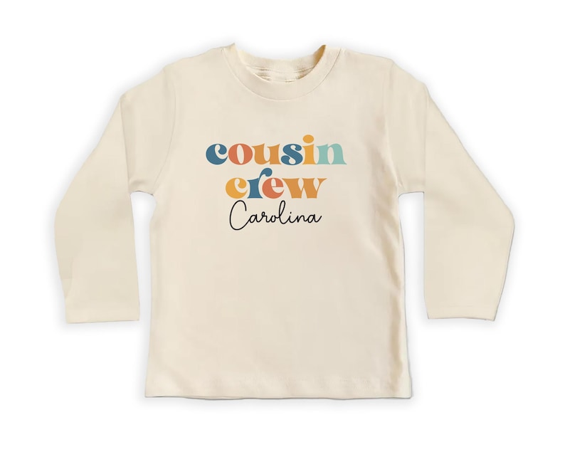 May include: A long-sleeved, cream-colored t-shirt with the text "cousin crew Carolina" in colorful retro-style lettering.