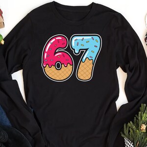 May include: Black long-sleeve shirt featuring a colorful graphic of the number 67. The number 6 is pink with red sprinkles and a waffle-cone base. The number 7 is light blue with sprinkles and a waffle-cone base. The shirt is a casual style.