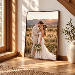 Puede incluir: Un cuadro rectangular con marco que muestra la fotografía de una pareja en un campo al atardecer. La persona de la izquierda viste un traje beige y la de la derecha un vestido de novia de encaje blanco mientras sostiene un ramo.