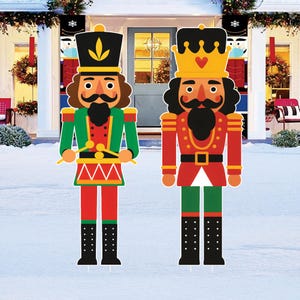 May include: Two large, colorful outdoor Christmas decorations of nutcrackers. One nutcracker is dressed in green and red with a black hat and holds drumsticks. The other is in red and gold with a crown. Both have black mustaches and boots.