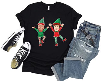 Może przedstawiać: Czarny T-shirt z grafiką dwóch kreskówkowych elfów w zielonych i czerwonych strojach elfów. Elfy uśmiechają się i mają uniesione ręce.
