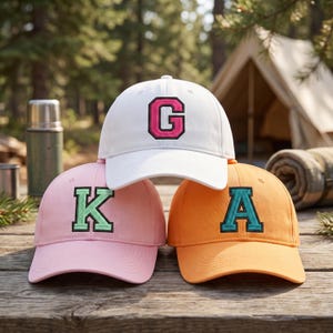 May include: Three baseball caps stacked together. The top white cap features a pink letter G with a black border. The bottom left light pink cap displays a green letter K with a black border. The bottom right orange cap shows a teal letter A.