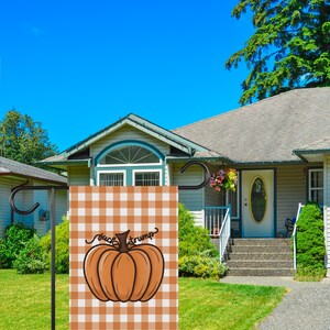 May include: A garden flag with a pumpkin design and the text "fuck trump" on a brown and white checkered background. The flag is displayed in front of a house with a blue sky.