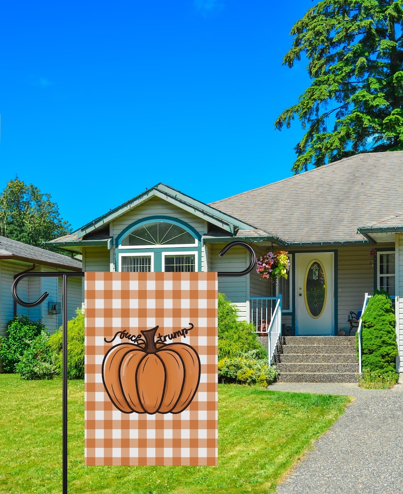 May include: A garden flag with a pumpkin design and the text "fuck trump" on a brown and white checkered background. The flag is displayed in front of a house with a blue sky.