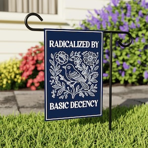 May include: A navy blue garden flag with the text "RADICALIZED BY BASIC DECENCY" in white. The flag features a white illustration of a bird perched on a branch surrounded by flowers and leaves. The flag is displayed on a black metal stand in a grassy garden.