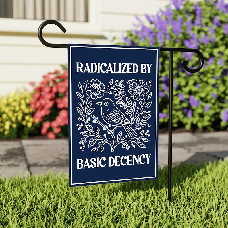 Puede incluir: Una bandera de jard&iacute;n azul marino con el texto "RADICALIZED BY BASIC DECENCY" en blanco. La bandera presenta una ilustraci&oacute;n blanca de un p&aacute;jaro posado en una rama rodeado de flores y hojas. La bandera se muestra en un soporte de metal negro.