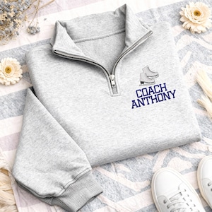 May include: A light gray quarter-zip sweatshirt featuring an embroidered graphic of a pair of ice skates above the text COACH ANTHONY in dark blue lettering. The garment has a high collar with a silver metal zipper and ribbed sleeve cuffs.
