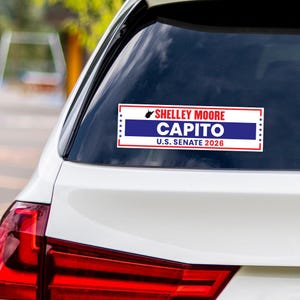 May include: A rectangular bumper sticker on a car's rear window. The sticker has a white background with a red border. It features the text "Shelley Moore Capito U.S. Senate 2026" in blue and white.