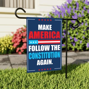 May include: A rectangular garden flag with the text "MAKE AMERICA FOLLOW THE CONSTITUTION AGAIN." The flag has a navy blue background with red and white text and stars. It is displayed on a black metal stand in a grassy garden.