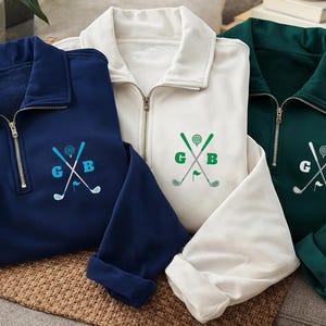 May include: Three golf-themed pullovers in navy blue, white, and dark green. Each pullover has a quarter-zip closure and embroidered golf club and ball design with the letters "G" and "B". The sleeves are cuffed.