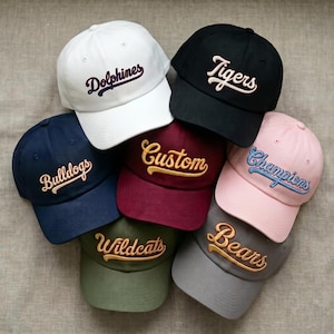 May include: A collection of seven baseball caps in various colours including white, black, navy blue, maroon, pink, olive green, and grey. Each cap features embroidered cursive text reading Dolphines, Tigers, Bulldogs, Custom, Champions, Wildcats, and Bears.