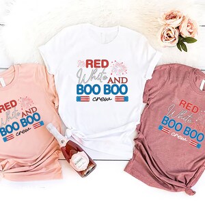May include: Three t-shirts with the text "Red White and Boo Boo Crew" in red, white, and blue lettering. The shirts are on a white wooden background.