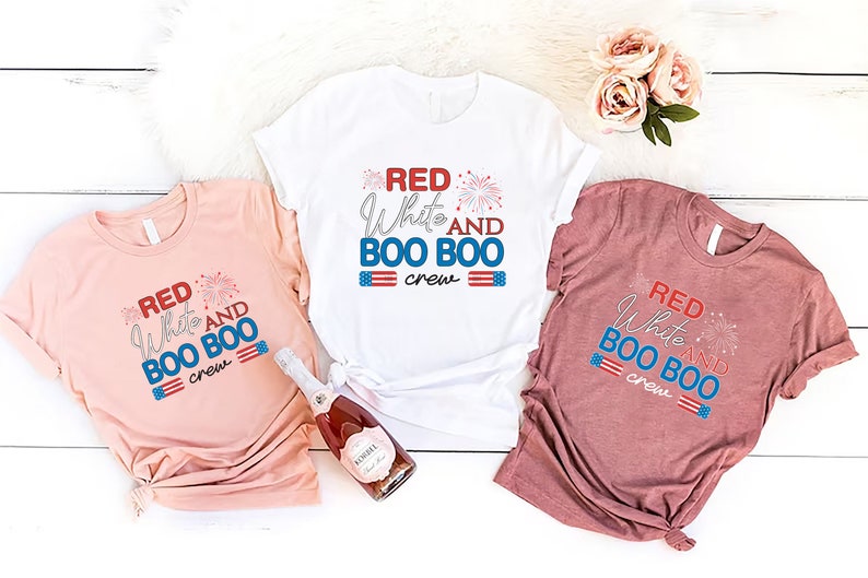 May include: Three t-shirts with the text "Red White and Boo Boo Crew" in red, white, and blue lettering. The shirts are on a white wooden background.