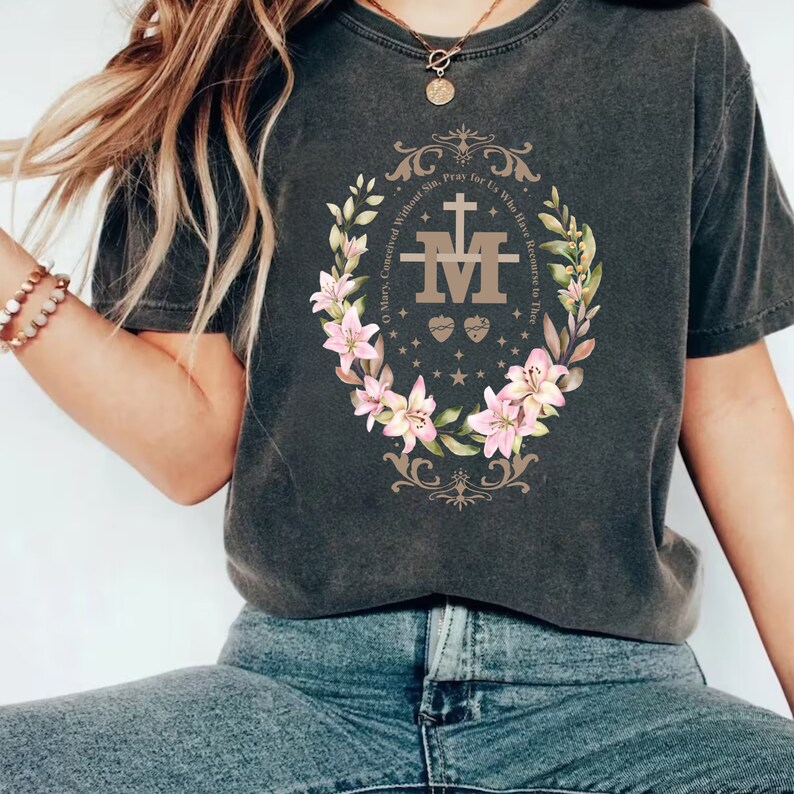 Op de afbeelding: Een donkergrijs T-shirt met een bloemenontwerp met een religieus symbool. De tekst op het shirt luidt "O Maria, ontvangen zonder zonde, bid voor ons, die onze toevlucht tot u nemen".