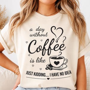 May include: A cream-colored t-shirt featuring black text that reads, a day without Coffee is like JUST KIDDING.... I HAVE NO IDEA. The design includes a graphic of a coffee cup with a heart on it, a heart-shaped steam swirl, and small hearts.