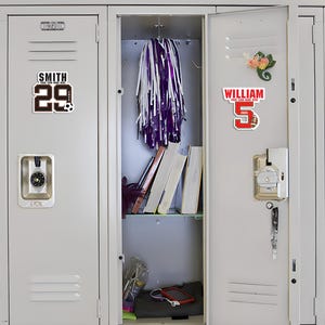 Peut inclure: Deux casiers m&eacute;talliques gris, l'un avec une plaque signal&eacute;tique sur le th&egrave;me du football qui dit "SMITH 29" et l'autre avec une plaque signal&eacute;tique sur le th&egrave;me du football qui dit "WILLIAM 5 2025". Les casiers sont ouverts, r&eacute;v&eacute;lant des livres, un pompon violet et blanc et d'autres articles.