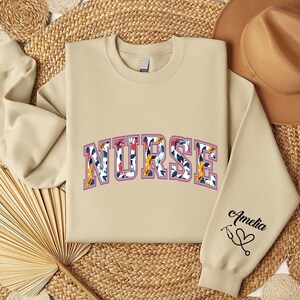 May include: A beige sweatshirt with the word "NURSE" in floral letters and the name "Amelia" with a heart on the sleeve.