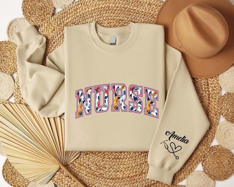 May include: A beige sweatshirt with the word "NURSE" in floral letters and the name "Amelia" with a heart on the sleeve.