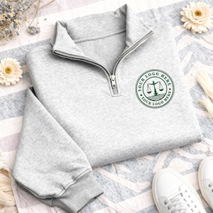 May include: A light gray quarter-zip sweatshirt with a silver zipper and a green circular logo that reads "YOUR LOGO HERE". The sweatshirt has a ribbed cuff and a folded collar. White sneakers are visible in the bottom right corner.