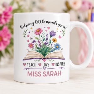 May include: A white ceramic mug featuring an open book with colorful wildflowers emerging from the pages. The text reads "helping little minds grow", "TEACH LOVE INSPIRE", and "MISS SARAH" in pink and black lettering with small hearts.