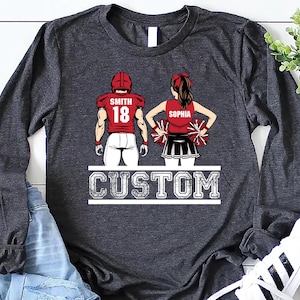 May include: Dark gray long-sleeve shirt featuring a football player and cheerleader design. The football player wears a red jersey with the name "Smith" and the number "18." The cheerleader's top says "Sophia." The word "CUSTOM" is printed below.