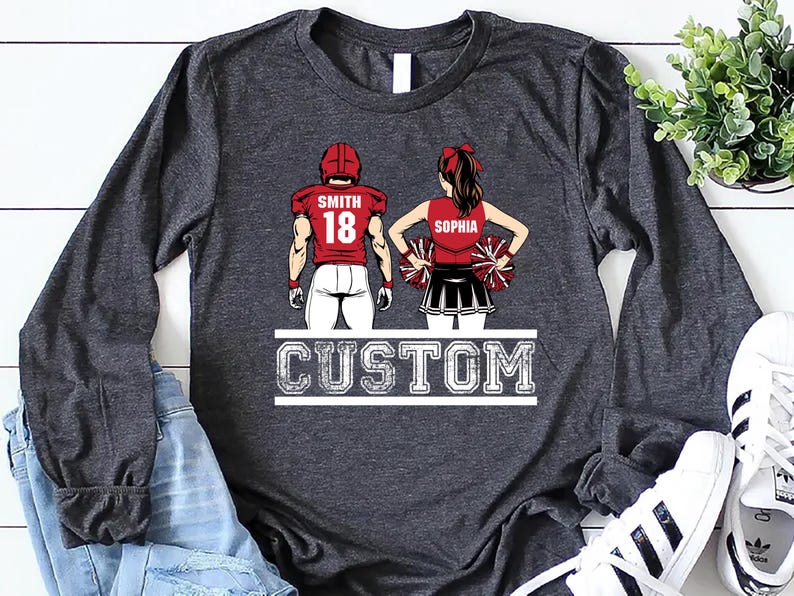May include: Dark gray long-sleeve shirt featuring a football player and cheerleader design. The football player wears a red jersey with the name "Smith" and the number "18." The cheerleader's top says "Sophia." The word "CUSTOM" is printed below.