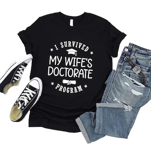 May include: Black t-shirt with white text that reads "I survived my wife's doctorate program". The text is surrounded by stars and includes a graduation cap and diploma graphic.